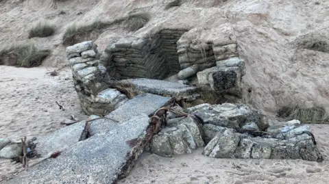 A grey concrete structure is poking out of the sand dunes. Sand bags filled with concrete create the walls of a hut-shaped structure. There are concrete slabs that appear to have collapsed in front of it. The structure is dotted with white splatters which may be lichens or as a result of weather damage.