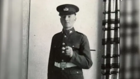 A black and white photo of a young Alan Davies wearing his military uniform and cap with a white cream belt. He is standing in front of a wall beside a floor-length curtain. He is looking at the camera with his baton neatly tucked under his arm. 