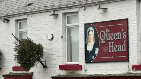 The Queen's Head Pub in Willington. The two-storey building is white, with a red door and windowsills.