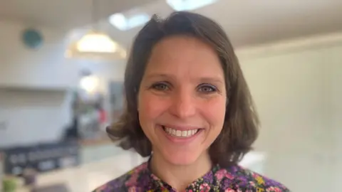 It's a close-up photo of a little brown-haired Eliza smiling at the camera and wearing a floral shirt. The background is blurred. 