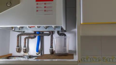 A boiler with rusty pipes coming out of the bottom and a bucket collecting dirty water on the right.