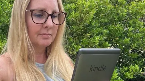 Kay, wearing a grey vest top and glasses and stood in front of a green bush, holds her 5th Generation Kindle in front of her
