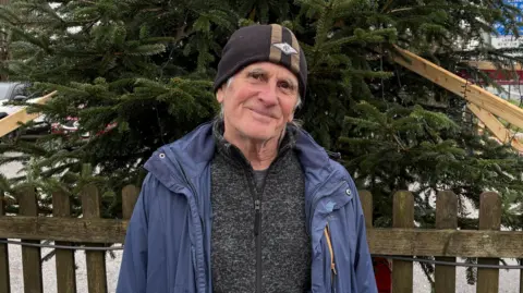 Roy wears a black beanie hat and dark blue raincoat and dark grey quarter-zip jumper. He is standing in front of a fence with a large Christmas tree immediately behind him.