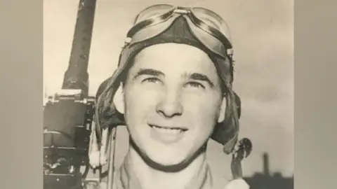 Casey Bukowski Casey Bukowski, pictured in black and white, wearing goggles on top of his head and holding a large gun over his shoulder. He is smiling.
