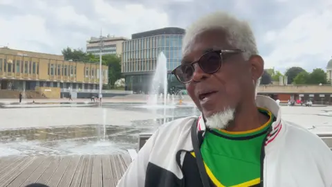 A man with white hair and a beard with sunglasses speaking outside in front of the mirror pool in Bradford