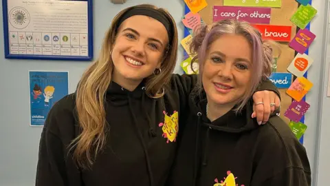 BBC / Ryan Dobney Ellie and Jan Hayes smile at the camera. They are both wearing black hoodies with a yellow and pink Starkidz logo on them. They are in a classroom.