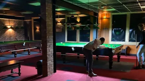 The Cue Club A man playing snooker - a dull room with red carpets and green snooker tables 
