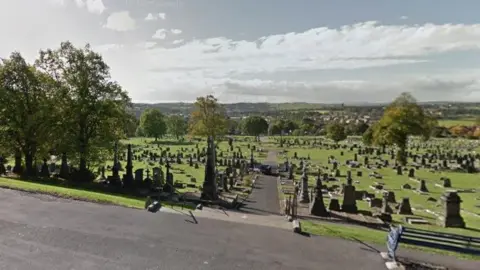 Google Dewsbury Cemetery with a path down the middle, grass and gravestones either side. The sky is sunny with clouds, and a bench is looking down the centre of the cemetery.