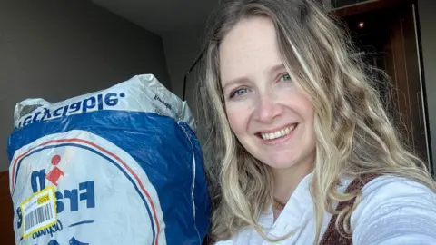 Photo of Laura who has long blonde hair and smiling. She is posing next to the bag of clothes she sent to avoid extra luggage costs. 