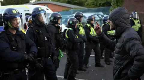 Reuters A man in a black jacket raises a hand and gestures at a line of riot police
