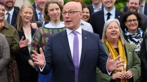 John Swinney, who is bald with glasses, poses in front of a cohort of SNP MSPs 