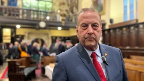 Phil Bialyk with a light blue shirt, a red tie and a mid-blue suit jacket with an Exeter badge on his lapel and a small microphone with the background of the Guildhall blurred