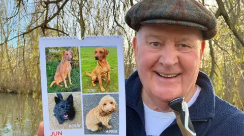 Tim Hart smiles as he holds up a large calendar. He is showing the page for June, which includes four photos of dogs. Mr Hart is standing in front of a canal and wearing a grey flat cap, a blue fleece and a white T-shirt.