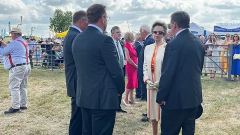 Princess Anne wearing a beige jacket and skirt talks to people at the show with stalls in the background.