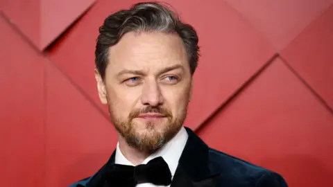 James McAvoy wearing a suit and a bow tie. There is a red background behind him. 