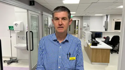 A man wearing a blue checked shirt is standing in the middle of a brand new emergency department. Behind him are hospital side rooms with doors and a desk area that will serve as a nurses station.