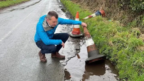 A man wearing a blue coat, black trousers and brown shoes picks up an orange and white traffic cone out of a pothole filled with water on the side of a road. There is a grass verge to his right.