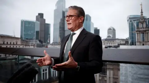 Keir Starmer standing and looking towards front-left corner of the image, talking and gesturing with his hands. He is standing on a balcony in London.