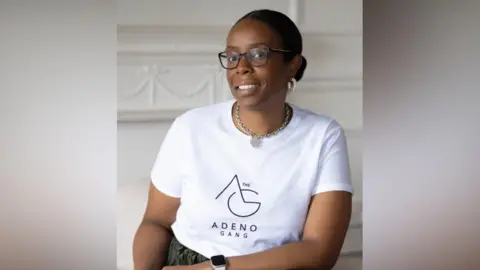Tanya Simon-Hall  is smiling at the camera, and is wearing a white branded t-shirt which read 'Adeno Gang'