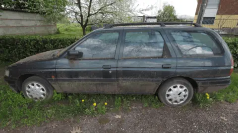 North Devon Council A black estate car with moss growing on the sides and bonnet is parked in  long grass next to a hedge.