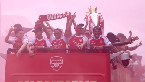 PA Media Arsenal players holding up their trophy and scarfs with the word 'Gunners' across them atop an open top bus looking happy watching onto the crowds on the streets. The air behind them is a misty red