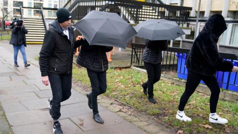 Four people arriving at court dressed in all black covered by umbrellas