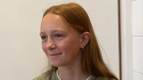 A girl called Ella. Has a straight brown hair which is long. She is wearing a green top. She has a gold ear-ring in her left ear.