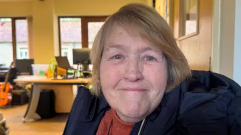 BBC/Emily Johnson An older woman sits indoors wearing a rust‑coloured sweater and a black hooded coat, with desks, computer monitors and a guitar in the background of a bright office.
