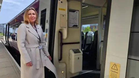 Philippa Cresswell with long blonde hair wearing a long beige coat. She has one leg on the step by the entrance to the coach of a train. She is standing on a train platform.