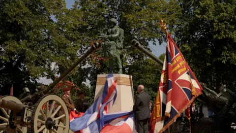 The statue is green and is standing on a plinth. There are cannons either side of it with flags adorning the surroundings. Trees are in the background.