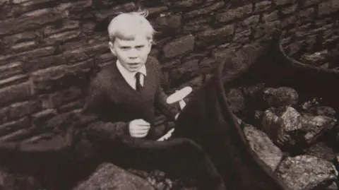 Getty Images Black and white picture of Jeff Edwards aged eight, with blonde hair, a dark jumper, white shirt and dark tie, holding a toy plane, looking at the camera, a few days after being rescued 