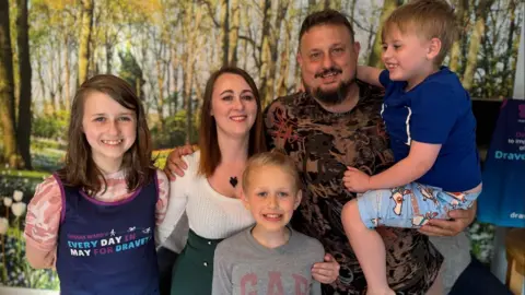 Lynda and Thomas Davies with their three children with woodland wallpaper behind them - all smiling. Lyidan is being carried by his dad Thomas.