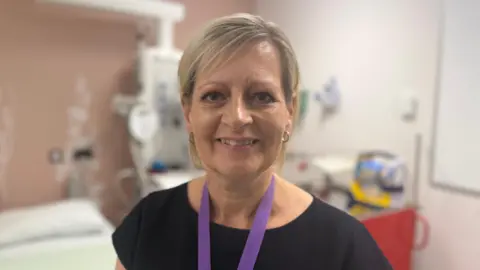 A lady with a blonde bob is smiling at the camera. She is wearing a black dress and a purple lanyard. She is standing in a hospital ward.