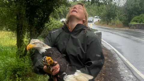 Sarah Hesford Sarah is standing beside a wet rural road, holding Aspen. She is wearing dark waterproof clothing.