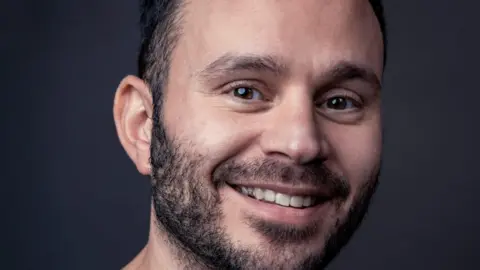 Nissen Lewis A professional headshot of a bearded man in his 30s smiling.