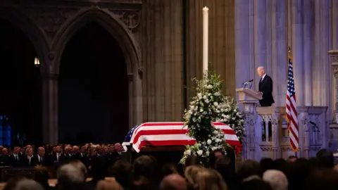 A coffin draped in a US flag lies in a packed cathedral with Joe Biden standing at a podium next to it.