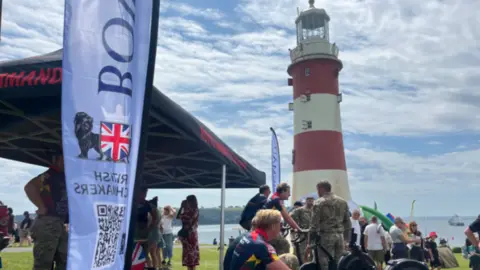 A group of people gather on the grass at Plymouth Hoe around military activity stands, with some using rowing machines and others chatting under a black canopy tent. In the background, a red and white lighthouse stands prominently against a bright sky, while flags, banners and more visitors fill the waterfront during the Armed Forces Day event.