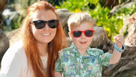 A woman with long red hair and sunglasses is smiling while crouching next to a little boy with blonde hair who is also wearing sunglasses and smiling. The boy has his fist raised in the air.