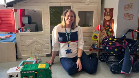 Clemmie Halligey sits in front of a child's Wendy House. She is wearing a white jumper with a navey blue stripe across it and navy blue trousers. She is surrounded by toys