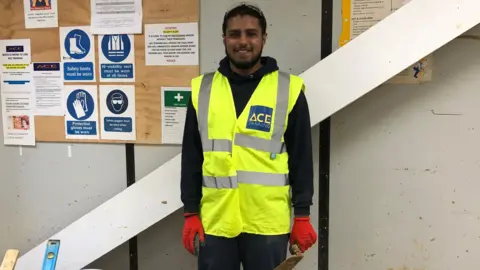 Muhammad stands in front of a wall with a wood noticeboard behind him. He is smiling into the camera and wears a black hoodie with a high-vis sleeveless jacket over the top. He is wearing red gloves and holds a trowel in  his left hand.