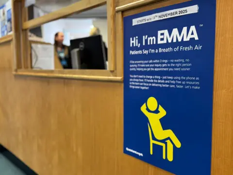 A blue poster featuring a yellow figure on a chair. The poster has large white text which reads "Hi, I'm Emma". It has been placed on a wooden wall. A reception desk is visible in the background and two people can be seen in the background.