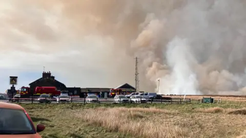Smoke fills the sky behind the Cat and Fiddle distillery. Fire engines are parked in the car park and a cordon is in place.