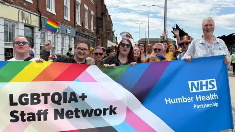 Four members hold out a colourful banner with LGBTQIA+ staff Network on - with the NHS Humber Health partnership logo on the right.