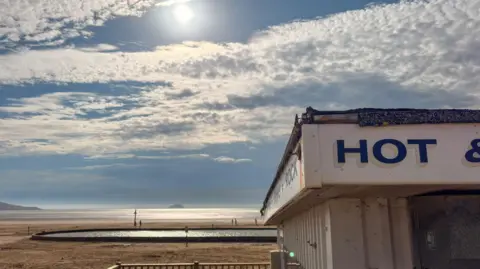 View of a beach with blue skies and sun. In the foreground is a food stall with the word Hot written in blue on the top.