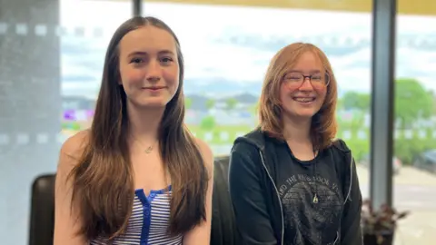 Two 16-year-old girls smile at the camera. They are sitting in front of a window.