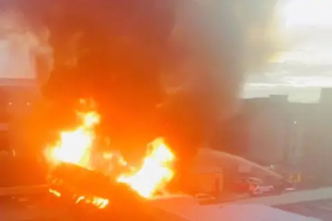 Innes Urquhart Flames in a building with smoke rising into the sky, with buildings in the background, and water from a hose going into the flames.