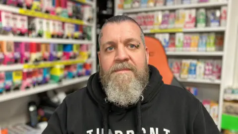 Brett Crundwell sitting in his vape shop in Didcot. He is wearing a black hoodie and has a grey beard. Behind him are brightly coloured vape packets.