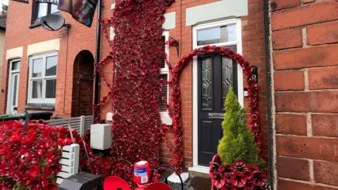 BBC A terraced house, with red poppies hanging from the top window. An arch is on display around the front door, and a Royal British Legion collection box can be seen on the front gate.