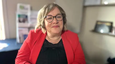 Martin Giles/BBC Jo Reeder is in an office room. she is wearing a red jacket and black top. She is wearing glasses and smiling. 