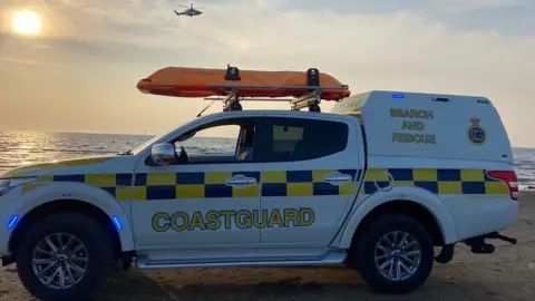 HM Coastguard Wirral A close up of the HM Coastguard Wirral's search and rescue vehicle parked on a beach near Leasowe Bay very close to the tide as the sun sets. A helicopter is in the air hovering near it.